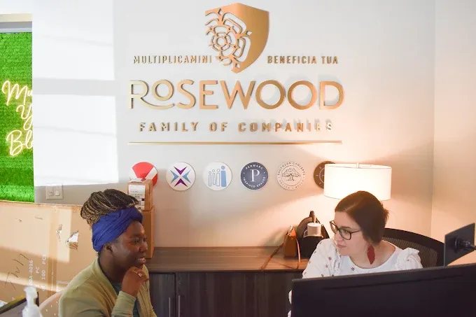 Two Rosewood team members talking at a desk beneath the Rosewood Family of Companies sign in the Raleigh office